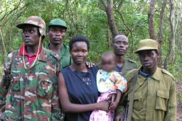 A woman is holding a baby, while four men in army fatigues stand beside and behind her. They are all standing in front of a forest, posed for the camera.