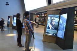 A man and woman stand looking at a double-sided video screen in a museum gallery. The man is pointing at the screen.
