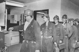 A line of miners hold lunch pails as they punch a time clock.