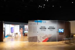 Upright panels are arranged in a circle in a dimly lit room. A gap offers people a way to enter the circle, and the interior of the circle is lit brightly. Next to the opening, a panel shows the words "Behind Racism: Challenging the Way We Think."