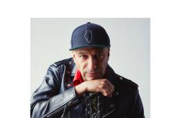 Tom Morello looks intently at the camera while leaning on his hand, which holds a guitar by the neck. He is wearing a black leather jacket, red collared shirt and baseball hat.