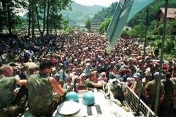 Two uniformed soldiers sit on a large vehicle, overlooking a densely packed crowd of people that stretches into the distance.