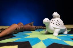 A white figure of a turtle with a frog on its back sitting on top of a colourful star blanket.