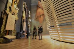 People in a Museum gallery explore tall wooden panels with wildlife and Métis beadwork. There are curved horizontal wooden slats to the right.