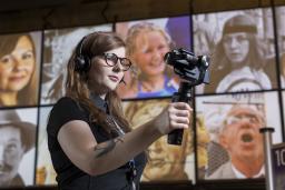 A woman wearing headphones, holding a mobile device on a handle, stands in a Museum gallery.