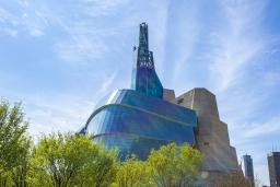 An unusual glass, steel and concrete building stands behind light green tree branches and green grass, against a bright blue sky.