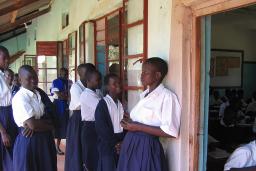 Un groupe de jeunes femmes et d’adolescentes debout sur la véranda d’un bâtiment. La plupart portent des blouses blanches et des jupes bleues, et deux filles portent aussi des chandails bleus. À droite, une porte mène vers une classe remplie d’élèves assises à des pupitres de bois et écrivant dans des carnets.