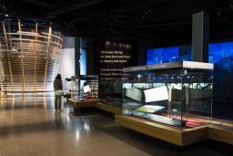 A museum gallery containing a large circular structure with horizontal wooden slats; glass cases containing artifacts; and projections on the wall.