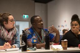 Three people sit at a table covered with water bottles, coffee cups and pads of paper.