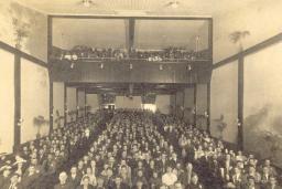  A black and white photo of a movie theatre audience. The picture is taken from the front of the theatre looking towards the back, so the faces of the audience can be seen. Potted palm trees line the walls on each side.