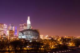 A city skyline at night. In the foreground is a river lined by trees, a glowing glass museum building and a bridge.