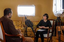 A man and a woman sit in chairs facing each other. There is a microphone on a stand positioned between them. There are also large lights on both sides of the woman’s chair, which are pointed towards the man.