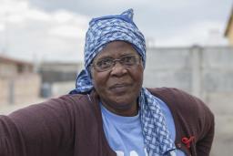 A head-and-shoulders portrait of Gogo Gladys Tyophol. She is wearing glasses and a blue patterned kerchief wrapped around her head. Her shirt says GAPA and a red AIDS ribbon is attached to her sweater.