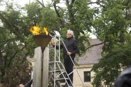 A man carrying a torch stands on a ladder to light a flame within a copper cauldron.