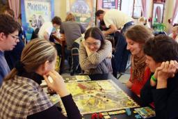 Three young women and two young men sit around a table playing a board game. There are other people playing board games at tables in the background.