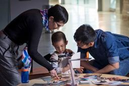 A woman, a man and a young boy are gathered around a circular table. Several photographs are scattered on the table, and the woman is pointing one out to the boy. 