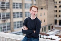 A man sits on the interior ledge of a rooftop. There are high-rise buildings in the background.