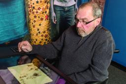 An older man sits in a wheelchair in front of a counter with a large touchscreen.