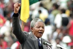 Nelson Mandela, an older dark-skinned man with short grey hair, stands in front of a podium and speaks into two microphones. He is wearing a grey suit with a tie. He is raising his right arm above his head and his hand is making a fist.