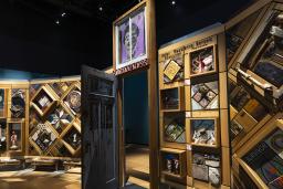 A large, curved freestanding wooden wall on which is mounted numerous small objects arranged in diamond and rectangular patterns. There is an open door in the centre of the wall.