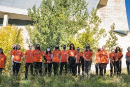 People wearing orange T-shirts stand in front of a row of trees. There is long grass in the foreground. The T-shirts bear images of small hand prints grouped in a heart shape with the number 215+ above them.