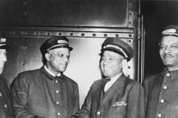 A black and white photo of four men in train porter uniforms. All of the men are smiling, and the two men in the middle appear to be shaking hands.