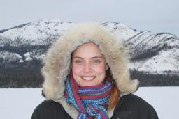 Smiling female wearing a fur-lined parka posing in front of a mountainous terrain.