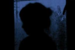 A dark image of a young child’s silhouette against a window through which are seen blurred trees and a power pole. The light is a purplish tone of twilight.