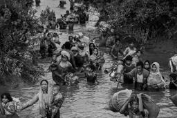Rohingya women, children and men wade through waist-high muddy river water. Some carry young children, while others carry bags of possessions, including household items.