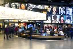 A large crowd in a museum gallery. There is a curved bench in the centre of the room. The display consists of a series of portraits above alcoves each containing a different exhibit.