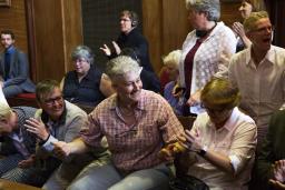 A group of people sitting on benches and standing in a dark wood panelled room. They are clapping and smiling.