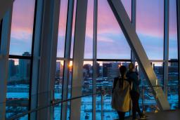 Two individuals stand looking through the floor-to-ceiling glass windows at the Winnipeg skyline. The sun is setting.