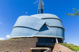 The Museum's entrance presents several ramp structures, some with grassy tops that extend all the way to the roof, which has a dome-shape formed by panes of glass resembling the folded wings of a dove. A tower rises in the center like a mountain peak. It is a sunny day with a bright blue sky and a small group of visitors are exiting the Museum.