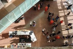 A bird’s-eye view of a crowd of people gathered in a large open area in the Museum. There are cocktail tables with red tablecloths and a bar on the left side of the image.