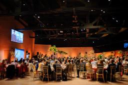 A crowd of people are seated around tables listening to a speaker in a large room.