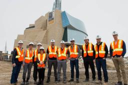 Ten people in hard hats and safety vests stand in front of a large, unique-looking building. The grounds are muddy and unfinished.