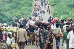 A large crowd of people of all ages carrying food and belongings walk toward the camera on a long dirt road through a bright green landscape of grass and bushes. The road and the crowd extend far into the distance.