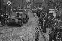 A crowd gathered on sidewalks, watching armoured vehicles driving through the cobblestone streets of a village.