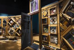 A large, free-standing art installation contains hundreds of objects. It is framed in cedar and there is an open door in the middle that people can walk through.
