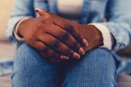 Close-up of a dark-skinned person sitting on a couch, with their hands clasped on their lap. 