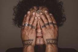 Aaju Peter covers her eyes and cheeks with her bare hands. Tattoos are visible on her forehead, chin, fingers and wrists. Her hands and the visible parts of her face are illuminated and are framed by her dark curly hair and angular shoulders.