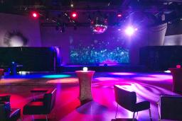 an empty, colorful event space with cocktail tables, lounge chairs, and a disco ball. The lighting creates a vibrant atmosphere, ready for a party or gathering.