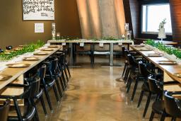 An open-concept bistro features a U-shape arrangement of long wooden tables decorated with green foliage, a white table runner and gold plates, and brown leather/wood chairs.