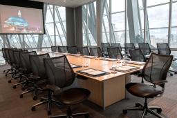 A boardroom enclosed by glass panels features a large screen, a long wooden table with notebooks and cups, and black mesh/leather chairs.