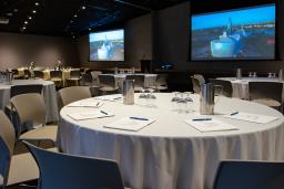 A large classroom is set up for a conference or, meeting with two large screens on the wall, round tables covered with white tablecloths, notepads and pens, and grey plastic chairs.