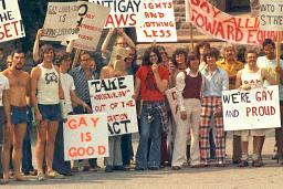 A large group of people dressed in T-shirts and seventies-style flared pants, stand together holding signs with messages such as “Out of the Closet,” “Gay is Good” and “Repeal Anti-Gay Laws.”