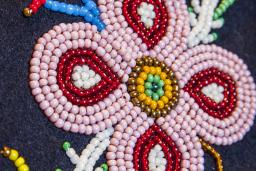 Close-up of a beaded floral design on dark fabric, featuring a pink four petal flower with red and white details, a yellow and green beaded center, and surrounding beadwork resembling leaves and stems.