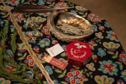 Smudging items arranged on a round table covered with a floral cloth, including an abalone shell holding dried sage, a feather, braided sweetgrass, cedar branches, and a small box of matches.