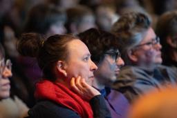 Audience members sit in a dimly lit venue watching a presentation, their faces illuminated by stage light. In the foreground, a person with their hair in a bun and wearing a bright red scarf rests their chin on their hand, looking attentively toward the stage. Other attendees sit beside them in soft focus, creating a quiet, contemplative atmosphere as the crowd listens.