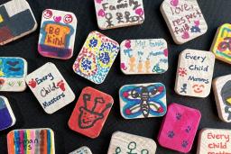 Small decorated wood tiles sit on a black backdrop. Some display images of families, others show messages like "Every child matters," "Be kind" and "Human is Human."